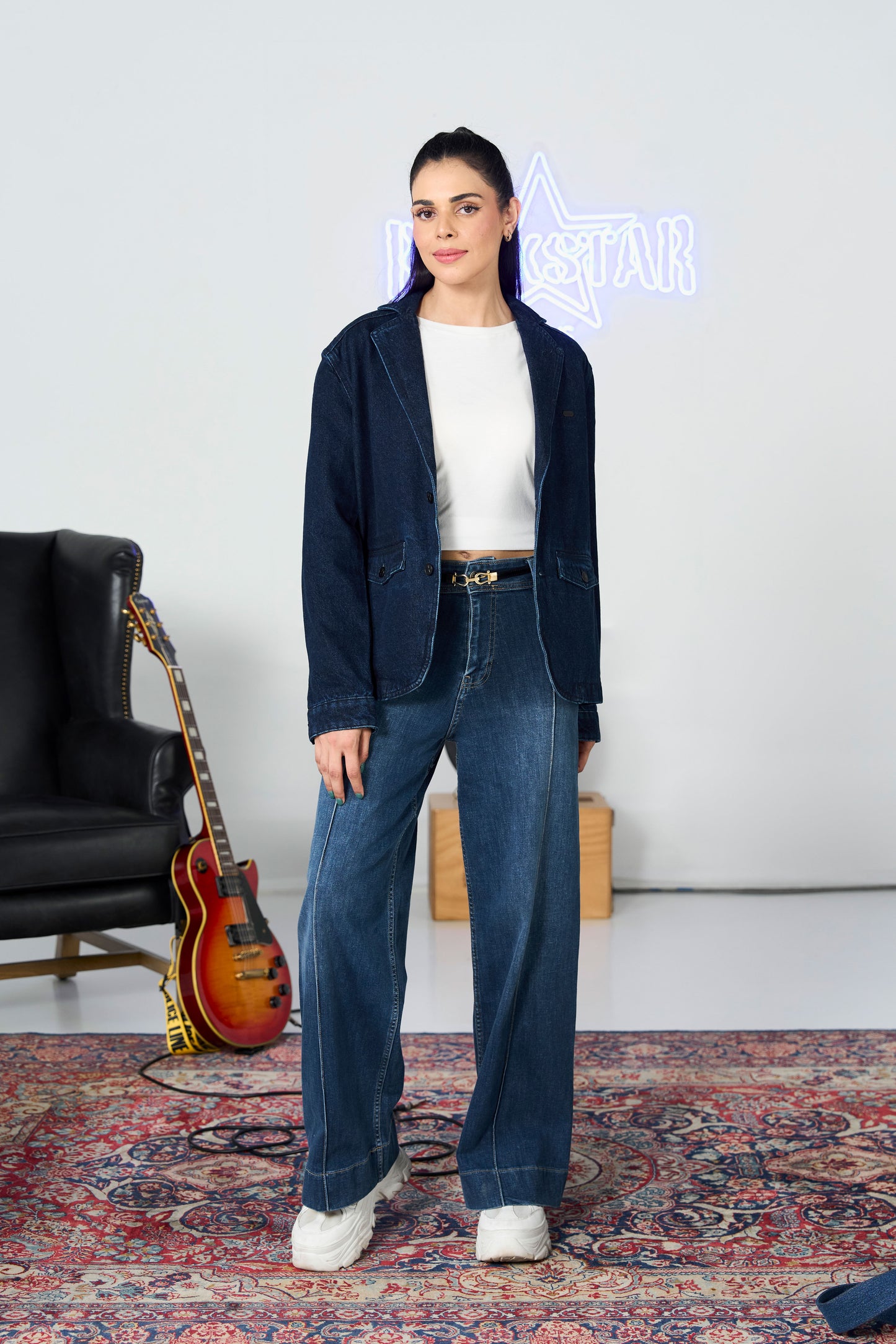 Woman in a navy blazer and blue jeans standing in a room with a guitar and neon sign.